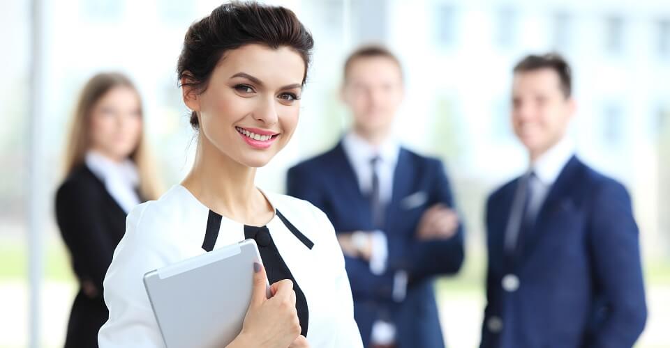 Business woman standing in foreground with a tablet in her hands, her co-workers discussing business matters in the background