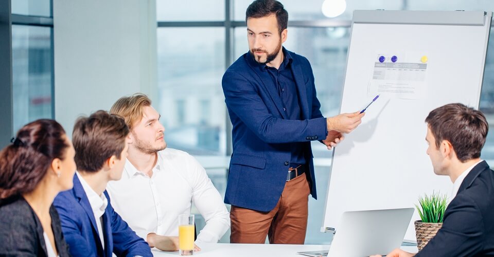 A man in a blue suit explains on the board to people sitting around a table